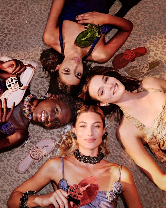 Four female models lying in a circle with their heads together on an animal print rug, holding sandals for Tory Burch.