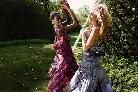 Two models in elegant dresses holding quilted chain handbags outdoors on a lush green lawn decorated with ornaments for Tory Burch holiday campaign