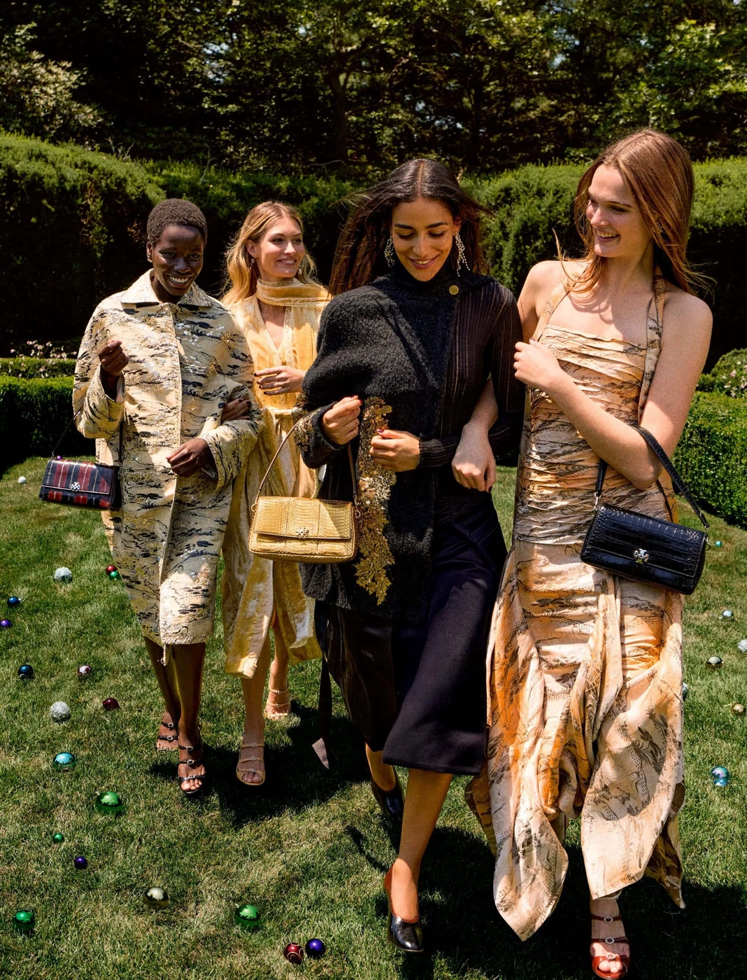 Four female models walking outdoors across a grassy lawn dotted with colorful ornaments, showcasing designer fashion for Tory Burch.