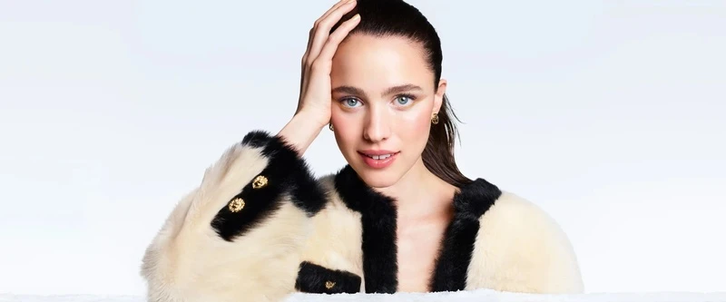 Margaret Qualley wearing a cream and black furry jacket with gold buttons, posing against a white background for Chanel.