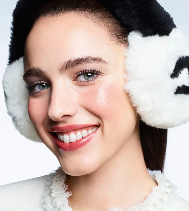 Model Margaret Qualley wearing fluffy white earmuffs and smiling for Chanel Holiday makeup campaign
