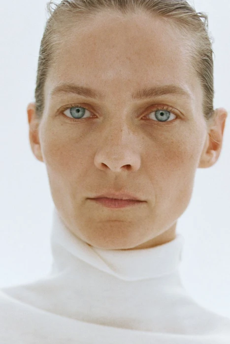 Close-up portrait of a woman with slicked-back hair wearing a white turtleneck against a plain background