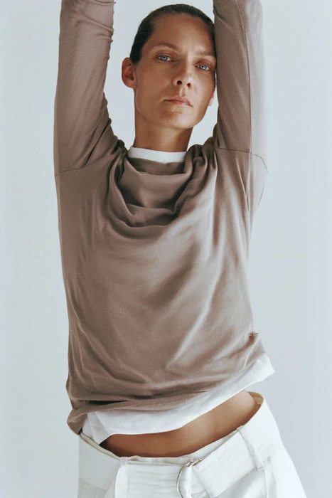 Model with arms raised wearing a brown long-sleeve top over a white shirt and light trousers against a white background