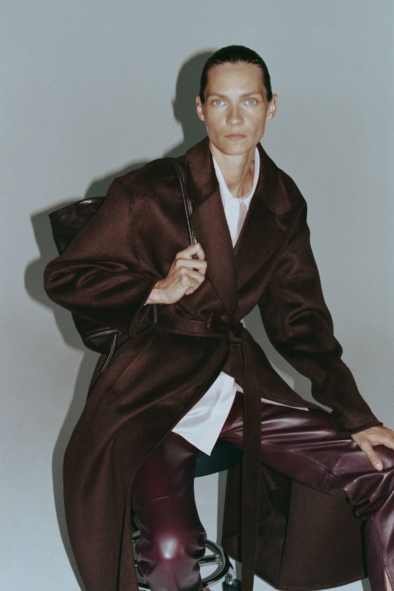 Model Karolin Wolter wearing a dark brown coat and leather pants with a black bag against a grey wall