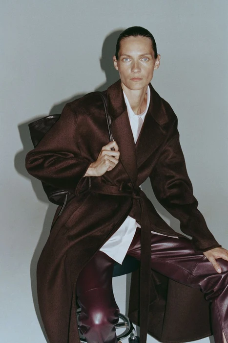 Model Karolin Wolter wearing a dark brown coat and leather pants with a black bag against a grey wall