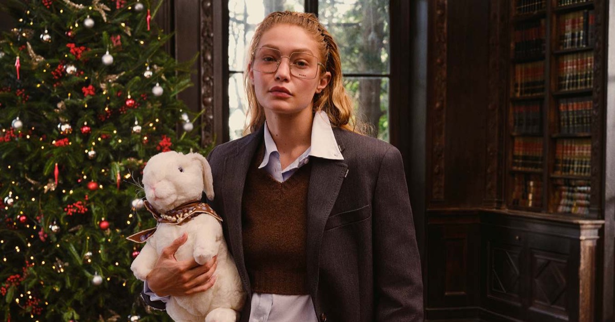 Gigi Hadid wearing large glasses and a dark blazer holding a plush white rabbit toy next to a decorated Christmas tree in a library.