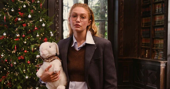 Gigi Hadid wearing large glasses and a dark blazer holding a plush white rabbit toy next to a decorated Christmas tree in a library.