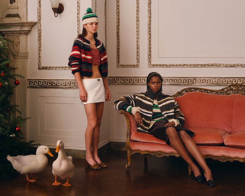 Two models posing in an ornate room with a Christmas tree and two white ducks on the polished wood floor.
