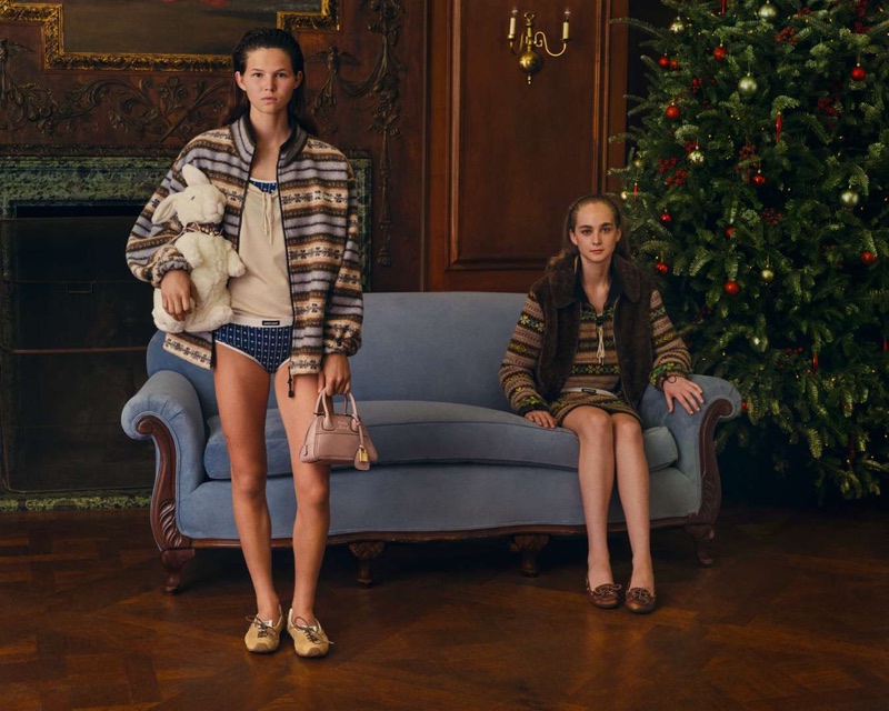 Two models posing in a warmly lit room with a blue sofa and decorated Christmas tree for the Miu Miu Holiday 2026 campaign.