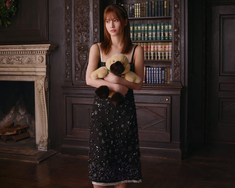 Woman wearing a black embellished slip dress holding a plush platypus toy inside a dark wood-paneled library room.
