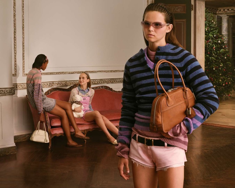 Model in striped sweater and pink shorts holding a brown leather handbag indoors with a red sofa in the background.