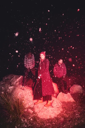 Three people stand on rocks in the dark with artificial snow. An adult woman in a red coat stands out in the center.
