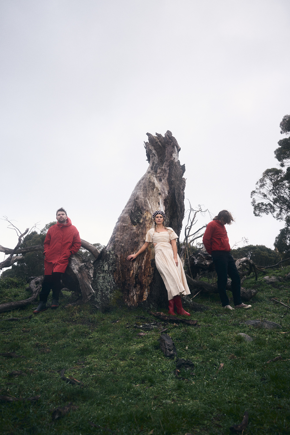 Trio stands near a massive fallen tree in a grassy, overcast field. Woman in dress and boots poses; others wear red jackets.