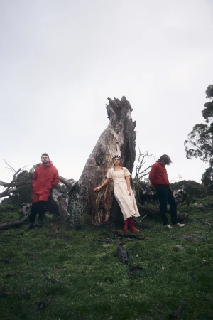 Trio stands near a massive fallen tree in a grassy, overcast field. Woman in dress and boots poses; others wear red jackets.