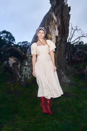 Woman in a cream dress and checkered hat poses near a tree on a grassy hill wearing red boots in a forest setting under a cloudy sky.