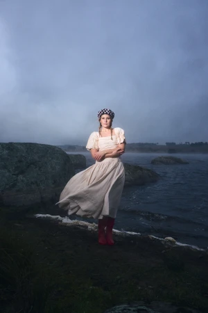 Woman wearing a cream dress and checkered beret standing on a rocky lakeshore under an overcast sky.