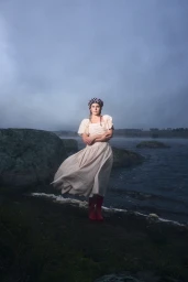 Woman wearing a cream dress and checkered beret standing on a rocky lakeshore under an overcast sky.