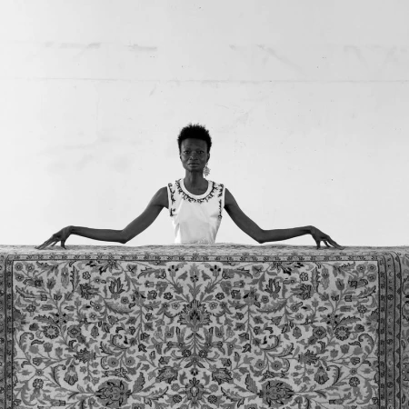 Black and white portrait of a Black woman behind a patterned rug, wearing an embroidered tank top and earrings, set against a white wall.