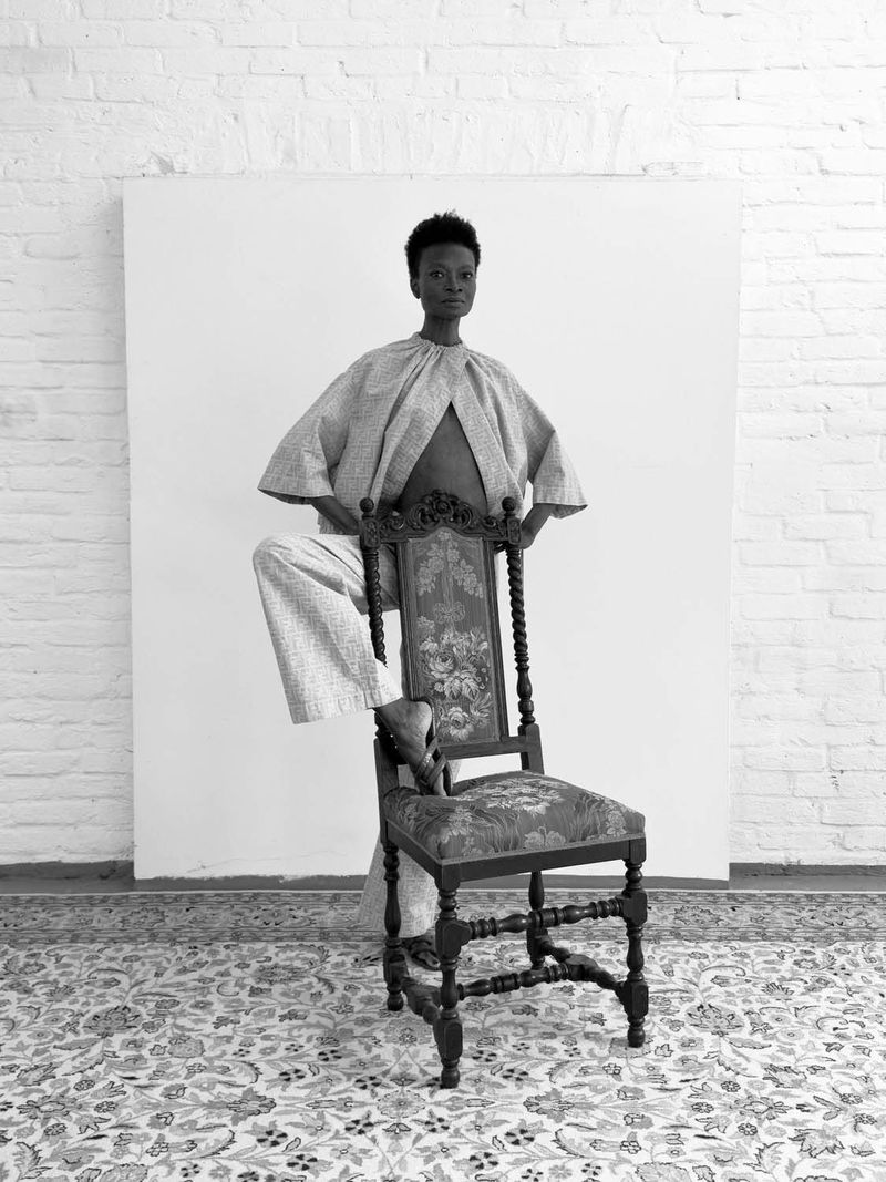 Monochrome portrait of a black woman in patterned separates, perched on an ornate vintage chair in a white studio with patterned carpet.