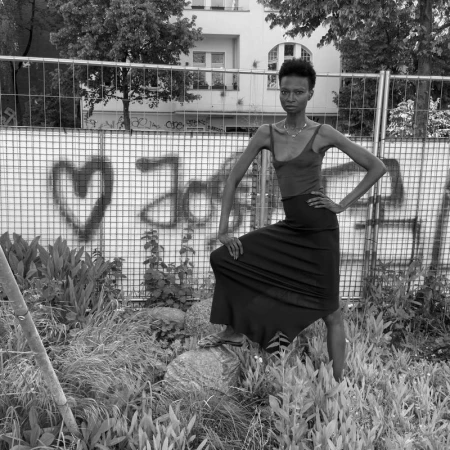 Black and white shot of an adult female in a skirt standing in a grassy lot in front of graffiti-covered fence near an urban building.