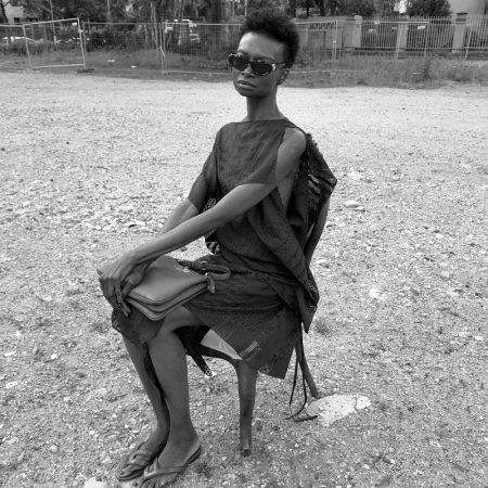 Monochrome portrait of an African-American fashion model in a black dress and sunglasses, seated outdoors with a handbag.