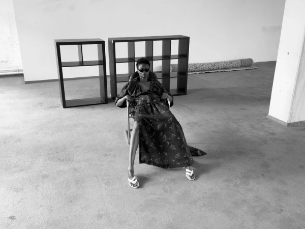 An adult female model in a long dress and sunglasses sits on a metal chair in a bright room, flanked by two black shelving units.