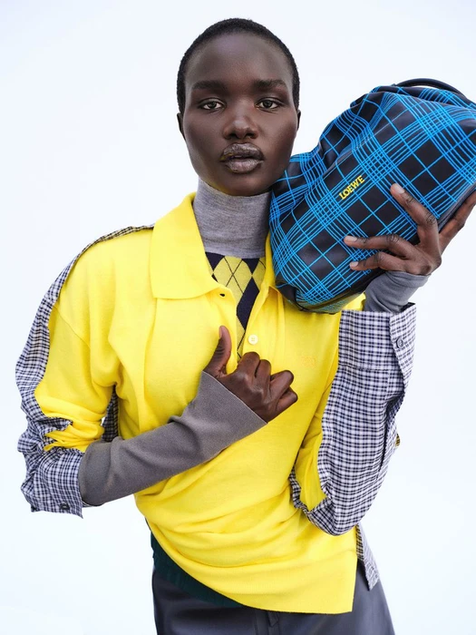 Model Akon Changkou holding a blue plaid Loewe bag wearing a yellow polo and grey turtleneck against a white seamless background