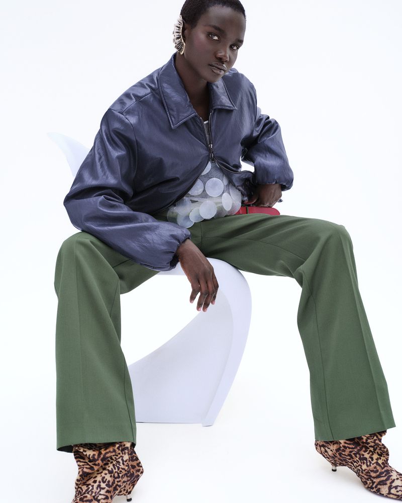 Model Akon Changkou seated on a white chair wearing a dark leather jacket, green trousers, and leopard print boots against a white backdrop.