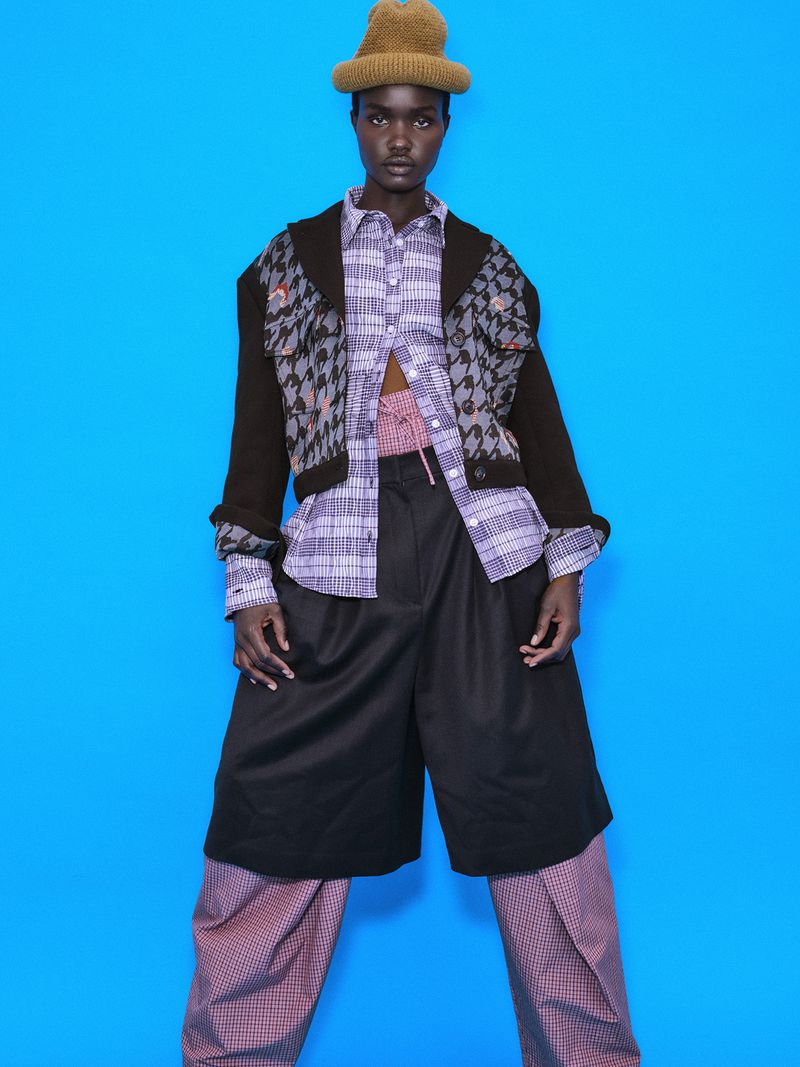 Akon Changkou wearing a patterned beanie, layered patterned shirt, dark jacket, and trousers against a vibrant blue studio backdrop