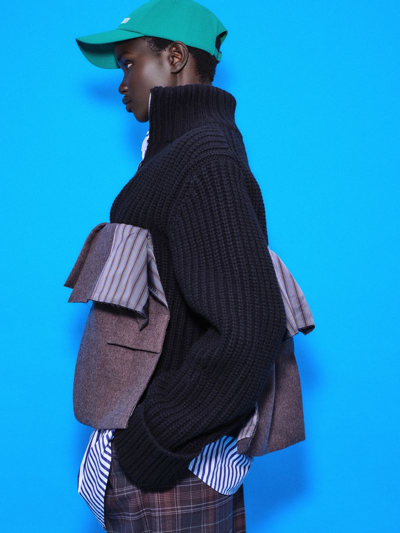 Model Akon Changkou wearing a green cap, black turtleneck sweater, and plaid trousers against a vibrant blue background.