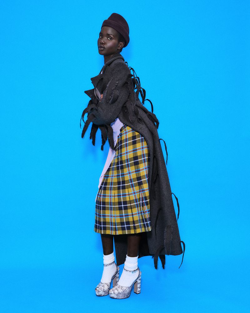 Model Akon Changkou wearing a dark fringed coat and plaid skirt against a vivid blue background