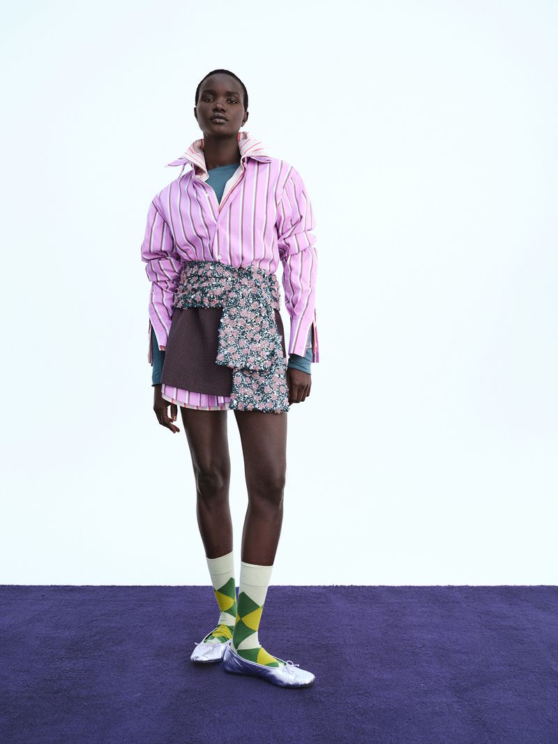 Akon Changkou modeling a striped pink shirt, brown skirt, patterned wrap, patterned socks, and silver flats against a white wall.
