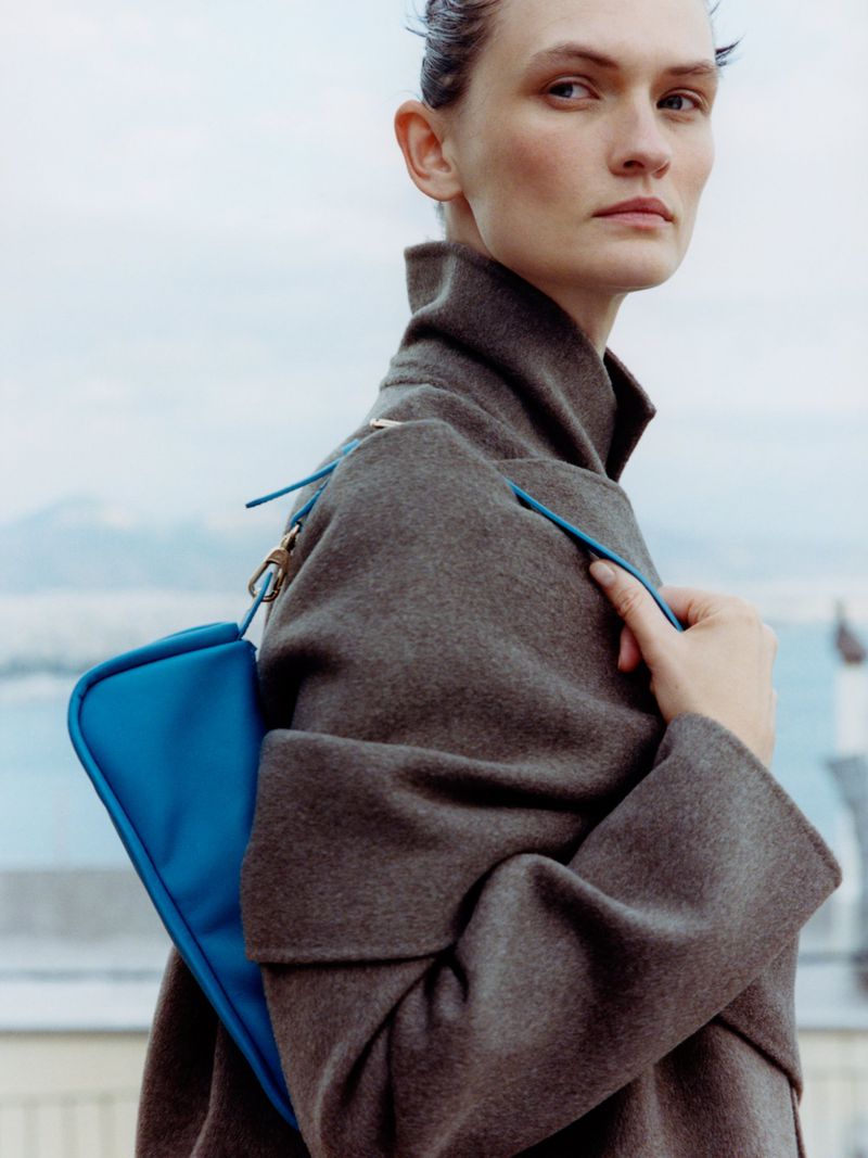 Model Lara Mullen wearing a grey coat and holding a blue handbag against an outdoor horizon for Vogue Greece