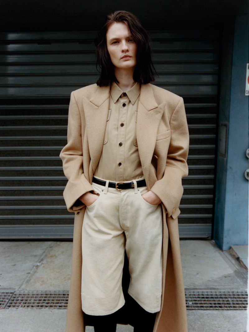 Model Lara Mullen wearing a camel coat and matching shirt against a dark corrugated shutter for Vogue Greece urban armor editorial