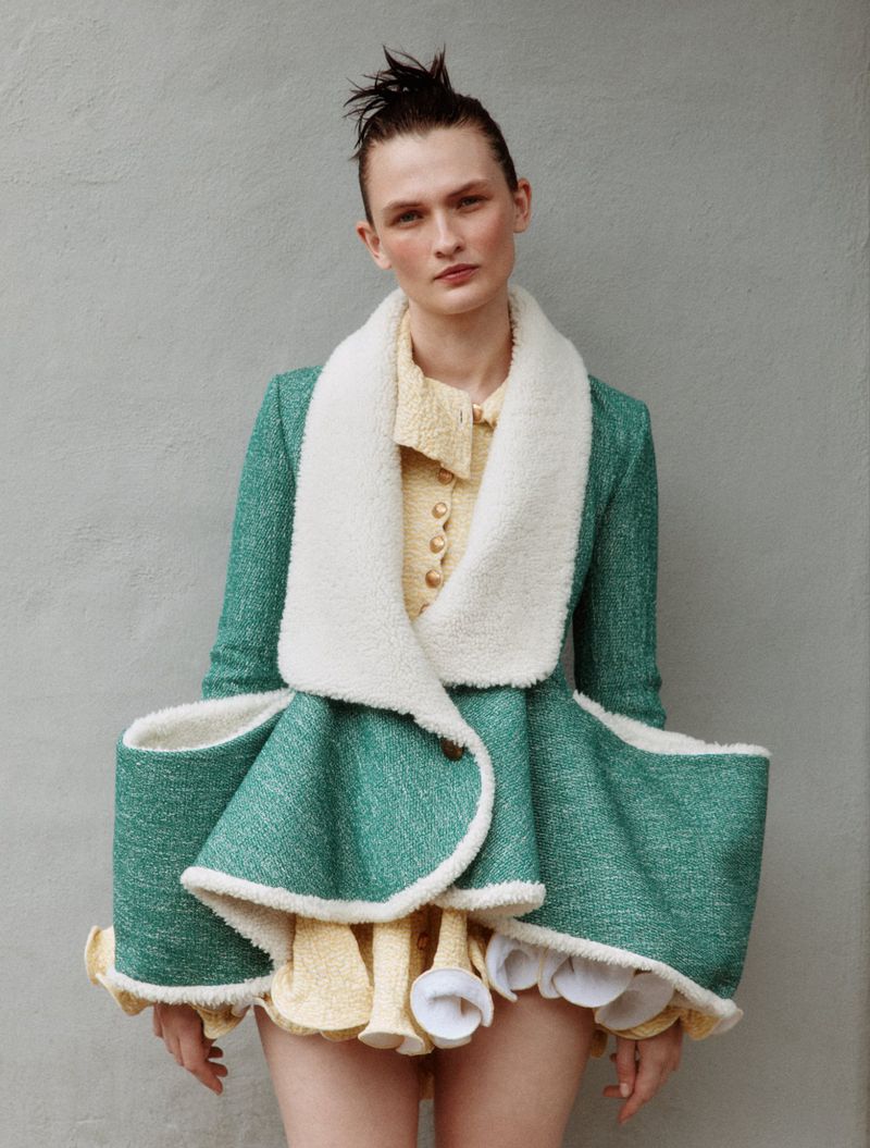 Model Lara Mullen wearing a structural green jacket with white trim and yellow dress against a plain textured background