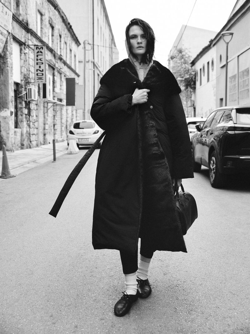 Model Lara Mullen wearing a long black coat holding a handbag on a street for Vogue Greece urban armor project