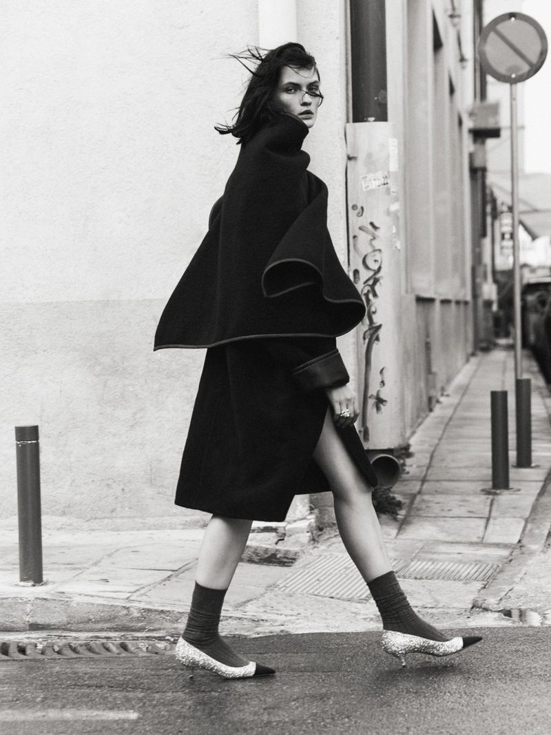 Model Lara Mullen walking down a city street wearing a black coat and sparkly heels for Vogue Greece