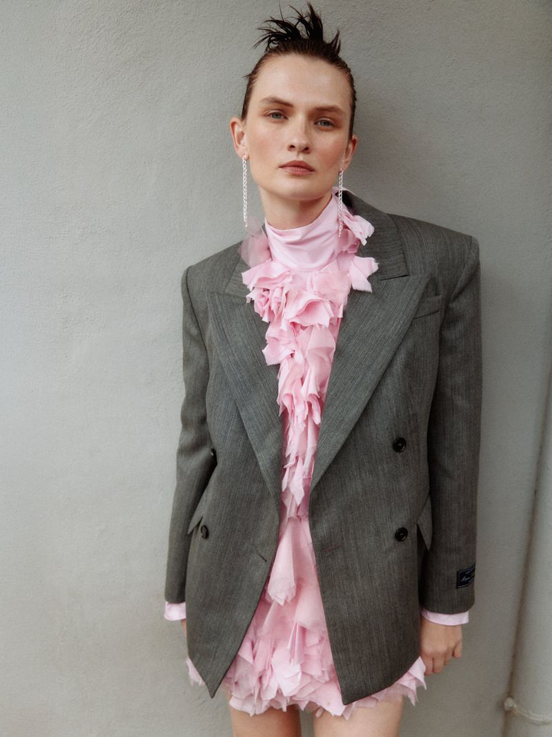 Model Lara Mullen in a grey blazer and pink ruffled top against a plain wall for Vogue Greece urban armor