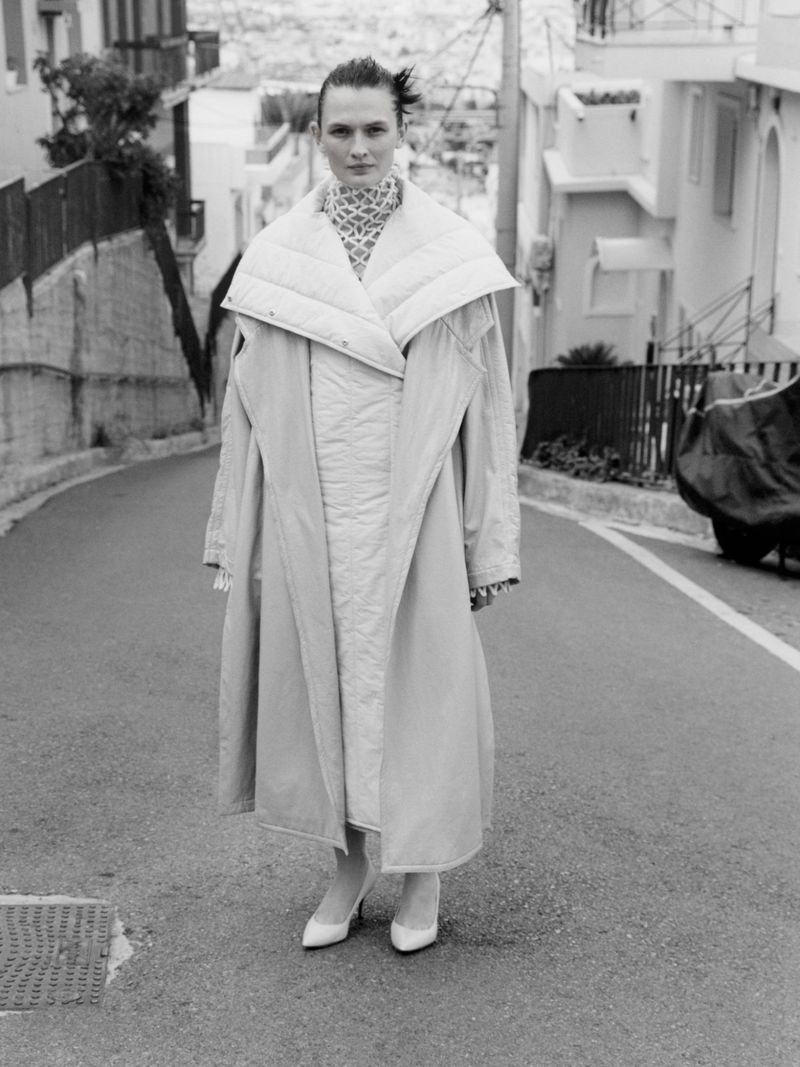 Model Lara Mullen wearing a quilted coat standing on a sloping street for Vogue Greece