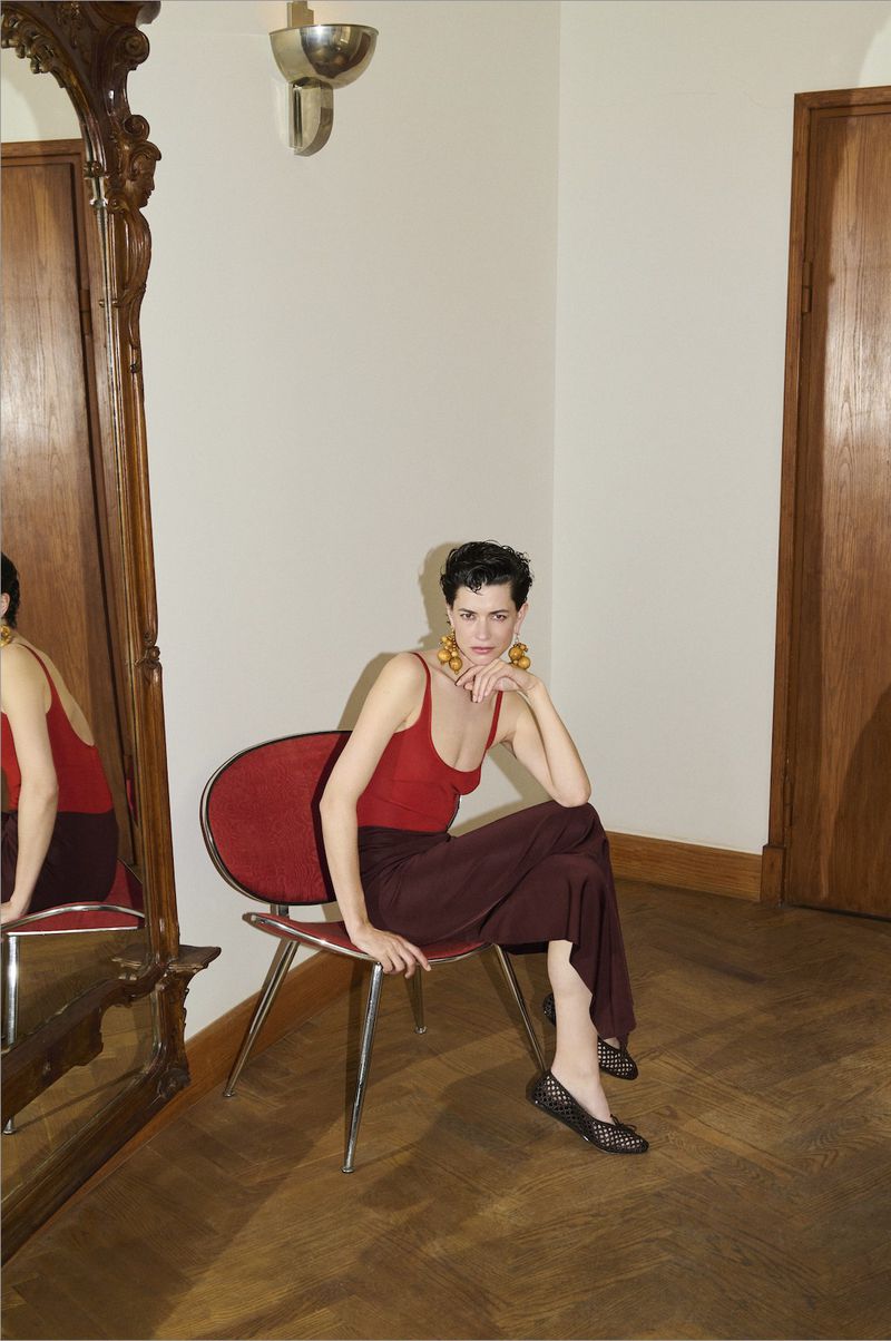 A stylish woman with short dark hair and gold earrings is sitting on a red chair in an elegant indoor setting with a large mirror.