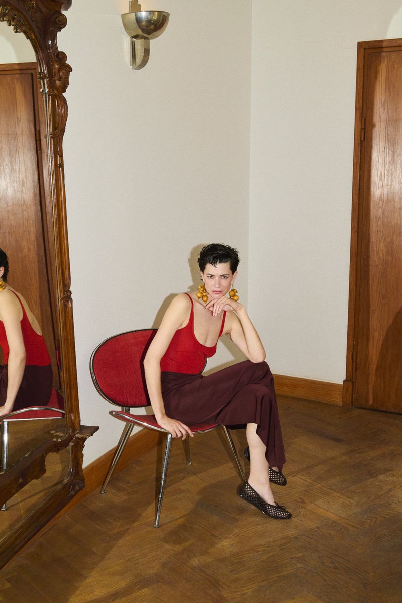 Woman in red top and maroon pants sits on a red chair near a mirror in an indoor setting with wooden floors and white walls.