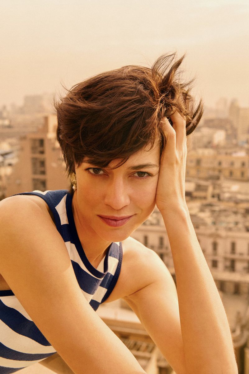 Portrait of a woman with short brown hair in a striped top, gazing at the camera with a city view as a backdrop.