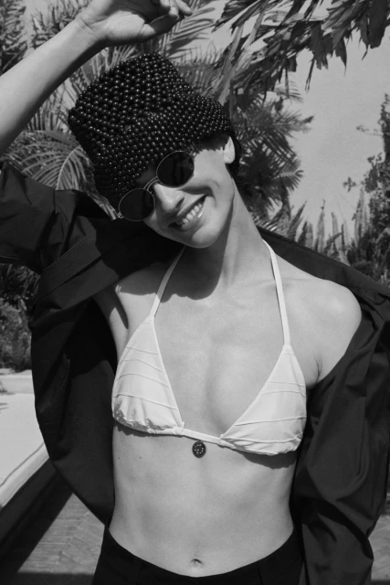 Black and white portrait of a woman wearing a beaded hat and sunglasses, smiling in a bikini top, with palm trees in the background.