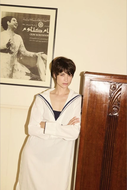 Woman in a white sailor-style dress poses indoors next to a vintage wooden cabinet, with a poster on the wall in the background.