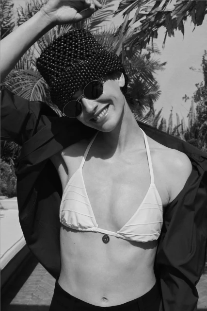 Black and white shot of an adult woman wearing a bikini top, beaded hat, round sunglasses and jacket posing in a tropical setting.