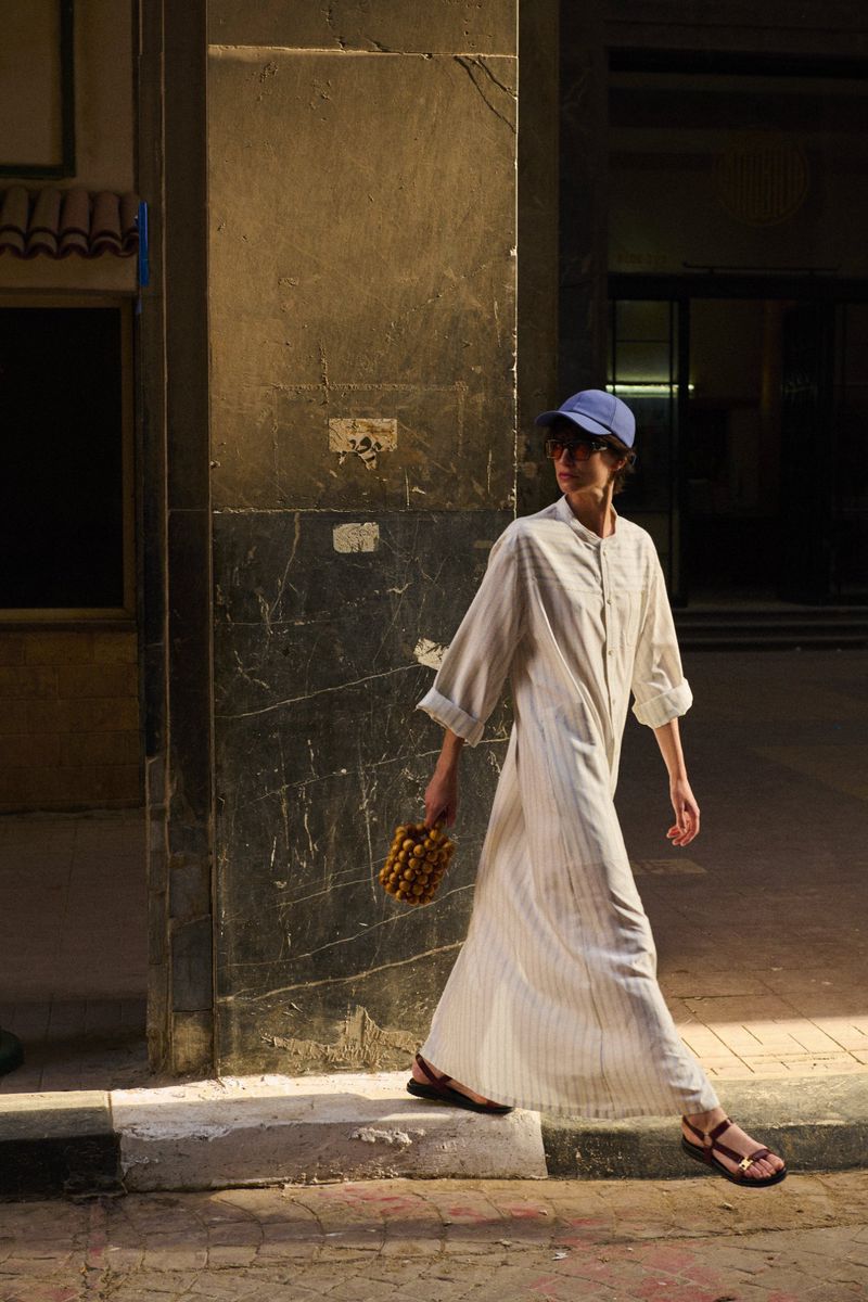 A woman in a caftan and cap walking along a city sidewalk carrying a beaded purse, illuminated by golden light from the side.