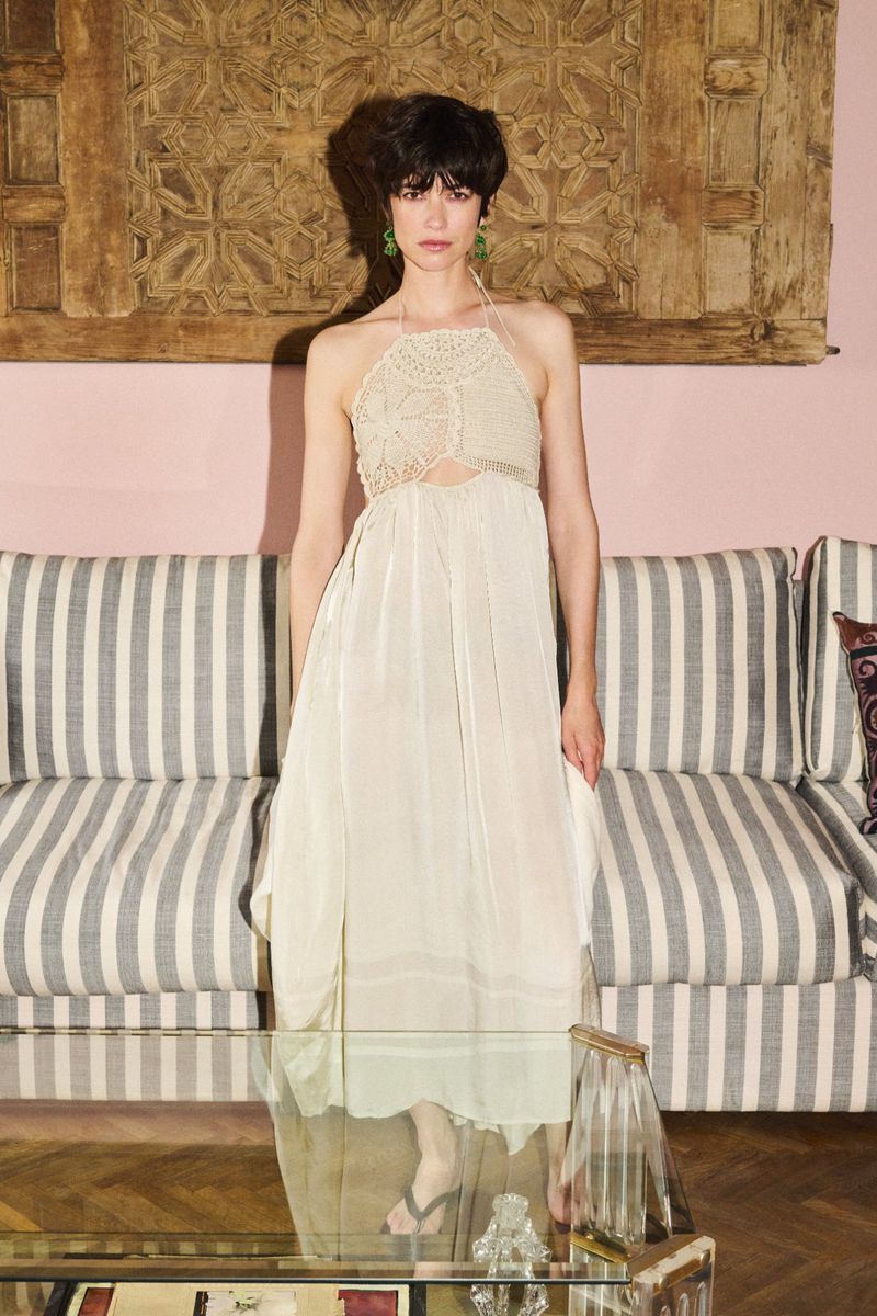 A stylish woman poses in a cream-colored halter dress against a vintage interior with striped couch and wooden paneling, exuding elegance.