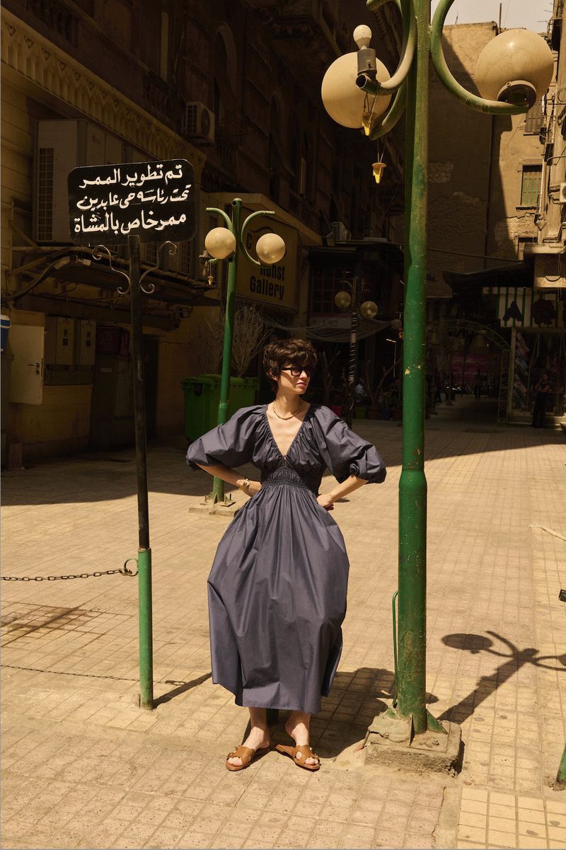 Woman in a blue dress poses between green lamp posts on a tiled sidewalk in an urban Middle Eastern environment during daylight hours.