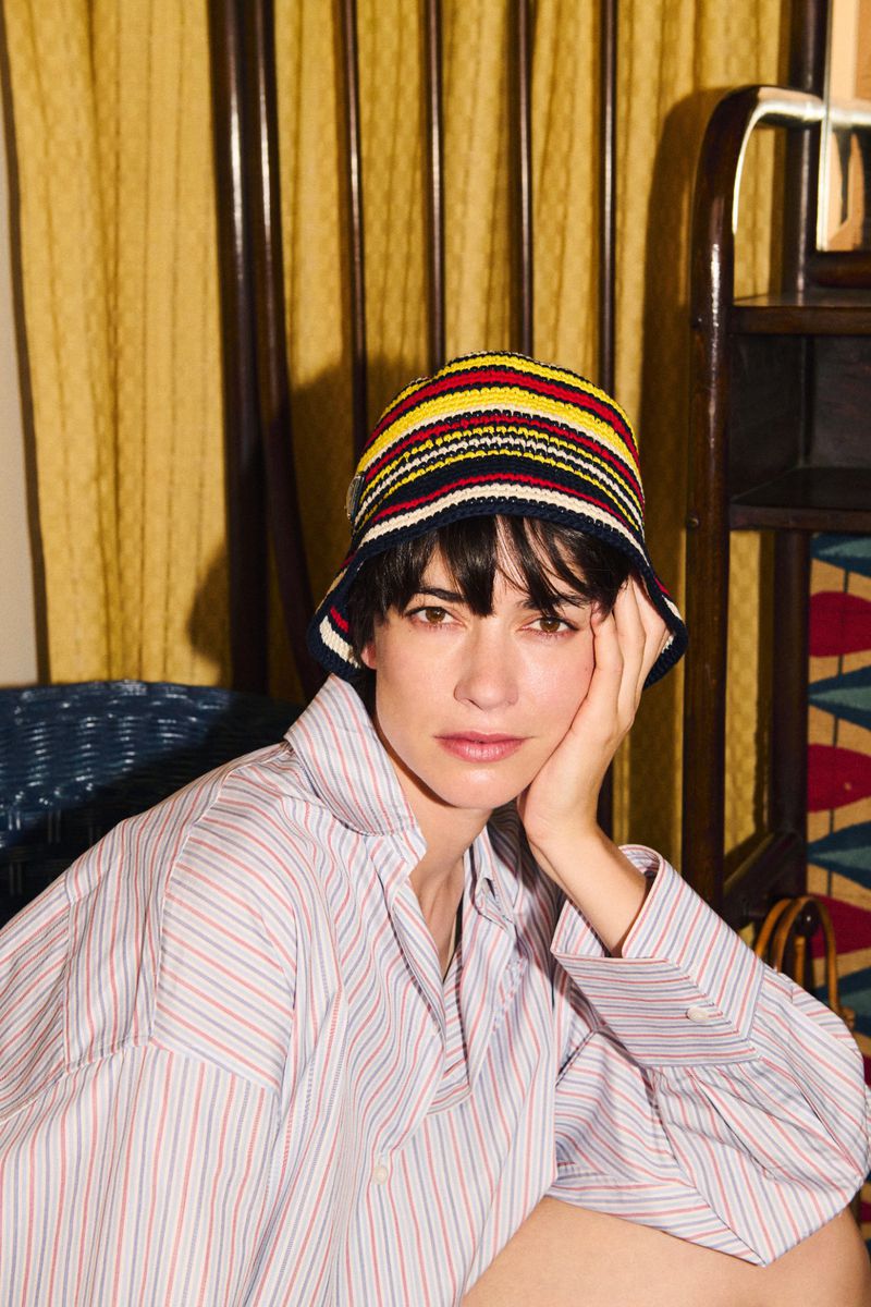 Portrait of an adult woman wearing a striped bucket hat and a striped shirt, indoors against a curtain and wood furniture.