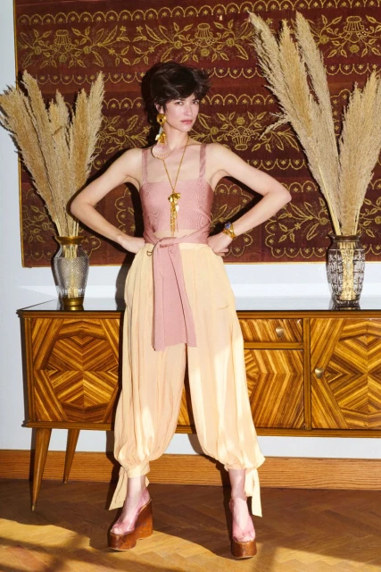 Indoor studio shot of a woman in a pink top and harem pants posing near a wooden cabinet and decorative wall hanging with pampas grass.